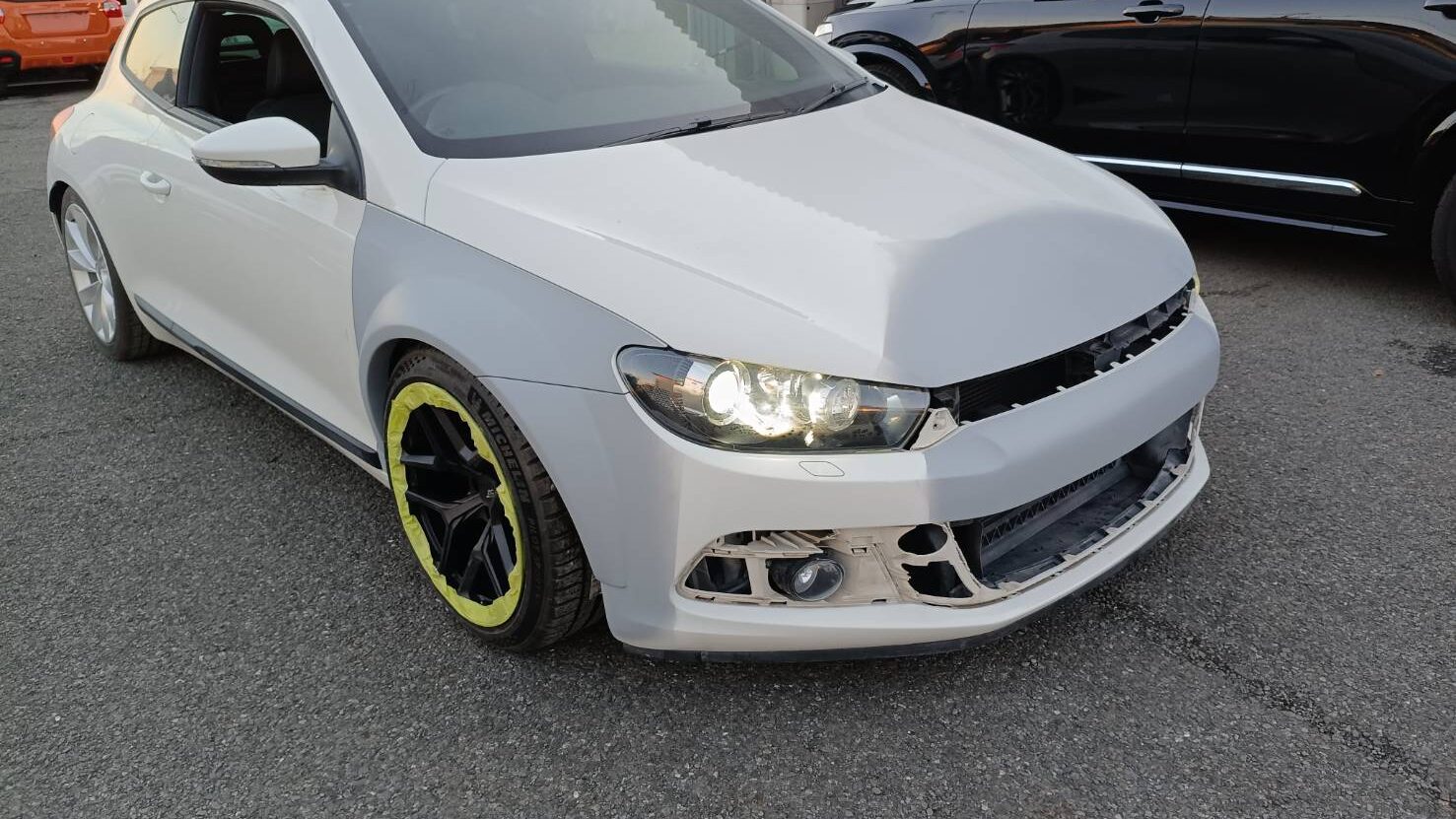 White two‑door car with gray primer on front fenders and missing front bumper, headlights on, black wheels with neon yellow rim tape.
