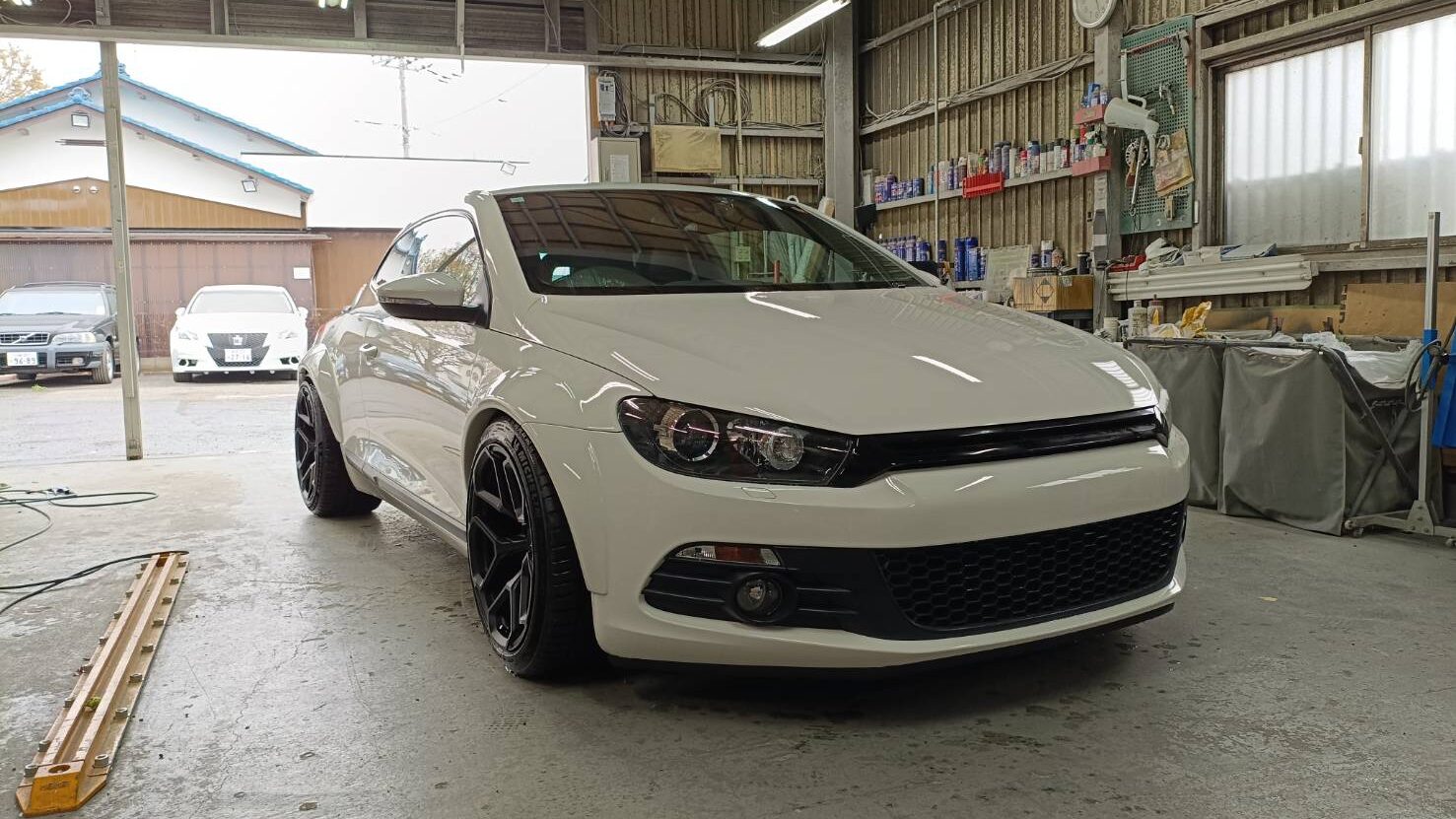 White modern sedan with black front grille parked inside a cluttered auto workshop, front-left view.