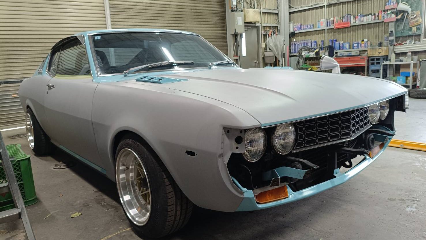Three-quarter front view of a vintage gray-blue car in a workshop, with the front bumper removed and teal trim visible on the hood and fenders