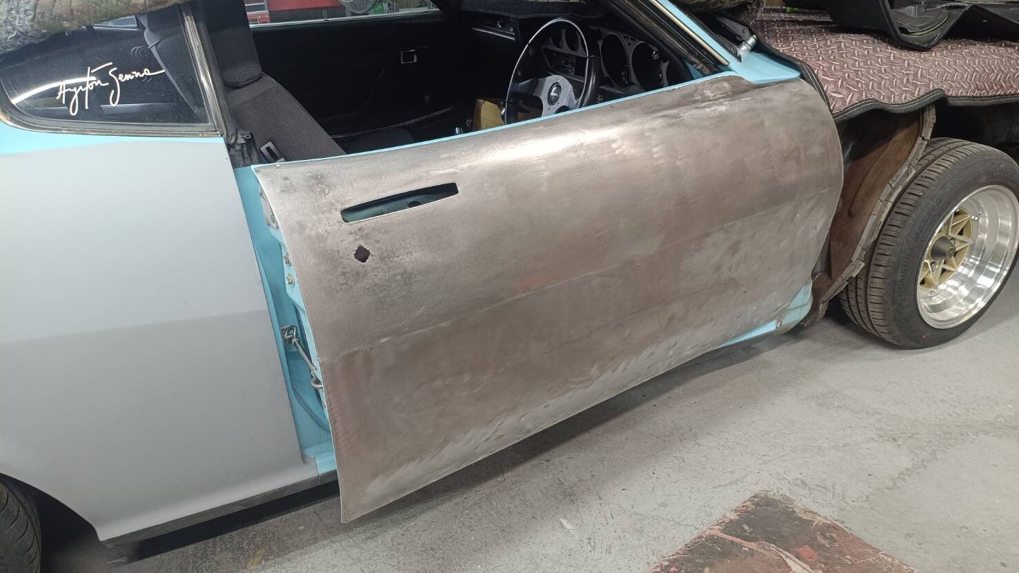 Vintage car in a shop with its door removed and a bare metal door skin resting against the side, exposing the interior.