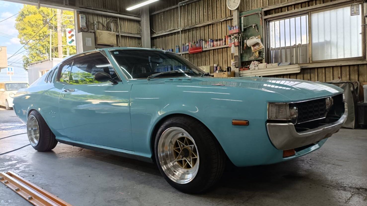 Light blue vintage two-door car in a cluttered workshop, chrome bumper and custom wheels visible, indoor garage setting